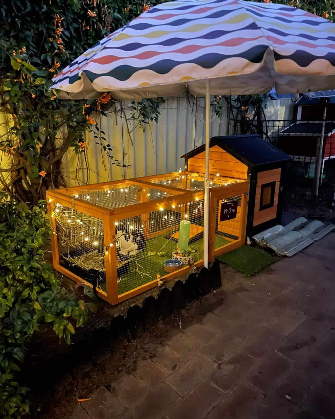 A brown and charcoal rabbit hutch decorated in fairy lights with an umbrella standing next to it.