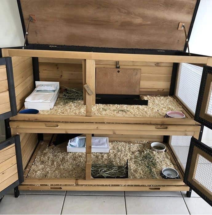 A two storey rabbit hutch with the doors open displaying hay bedding and food and water bowls.